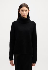 Melna turtlenecka džemperis ar teksturētu adījumu, ar ribotām aprocēm un brīvu piegriezumu, apvienots ar melnu svārku, minimālisma dizains.
