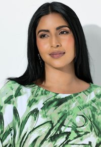 Zielony top z wzorem kwiatowym, z krótkimi rękawami, okrągłym dekoltem i subtelnymi zdobieniami. Długie ciemne włosy i neutralna pomadka.