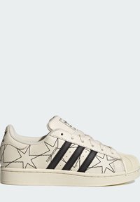 Baskets en cuir blanc avec motifs d'étoiles noires, trois bandes noires, embout en caoutchouc texturé et fermeture à lacets. Marquage "SUPERSTAR" sur le côté.