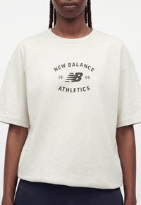 Άτομο που φοράει ένα ανοιχτό μπεζ t-shirt New Balance Athletics με μαύρο λογότυπο και κείμενο, σε συνδυασμό με σκούρο παντελόνι, μπροστά σε λευκό φόντο.