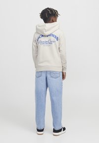 Felpa beige con una grafica blu che recita "Jack & Jones Denim Crew Collective" sulla schiena, abbinata a jeans baggy blu chiaro e scarpe nere.