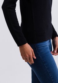 Schwarzes Langarmoberteil mit tailliertem Design und weicher Textur, kombiniert mit blauen Jeans. Das Outfit hat ein dezentes Saumdetail.