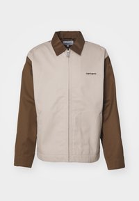 Lichtbeige en donkerbruine jas met een rits aan de voorkant, contrasterende mouwen en een Carhartt-logo op de borst. Gemaakt van gladde, duurzame stof.