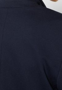 Námořnická modrá bunda s hladkou texturou a strukturovaným límcem, která má jemný švový detail na zadní straně. Klasický design a střih.