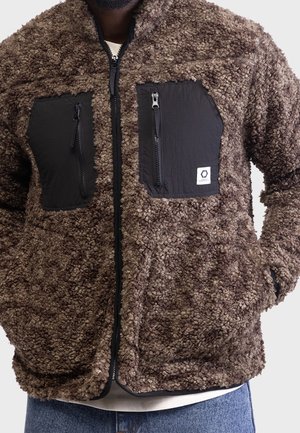 Mann trägt braune, strukturierte Fleecejacke mit schwarzen Reißverschlusstaschen auf der Brust und schwarzem Frontreißverschluss, Hände in den Seitentaschen, blaue Jeans sichtbar.