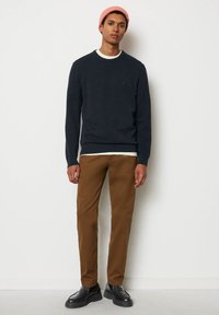 Dunkelblauer Pullover mit Rundhalsausschnitt, über einem weißen Hemd getragen; kombiniert mit braunen Chinos und schwarzen Schuhen; trägt eine rosa Strickmütze.