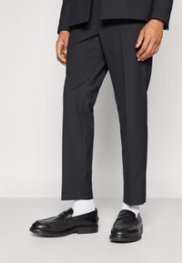 Schwarze Leder-Loafers mit glatter Oberfläche und dekorativer Naht, getragen mit weißen Socken und einer schmal geschnittenen schwarzen Hose.