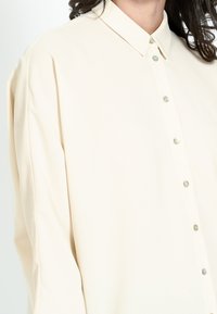 Krems krāsas krekls ar pogām un klasisku apkakli, izgatavots no gludas auduma, ar smalku spīdumu un noapaļotām pogām pa priekšu.