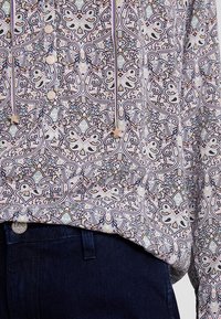 Bluzka z długim rękawem z lekkiego materiału, złożona z skomplikowanego wzoru kwiatowego w kolorach fioletowym, niebieskim i kremowym. Zawiera guziki oraz sznurki.