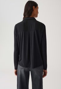 Camicia nera a maniche lunghe con una texture liscia, caratterizzata da un pannello a contrasto nella parte superiore e un sottile dettaglio plissettato sul retro.