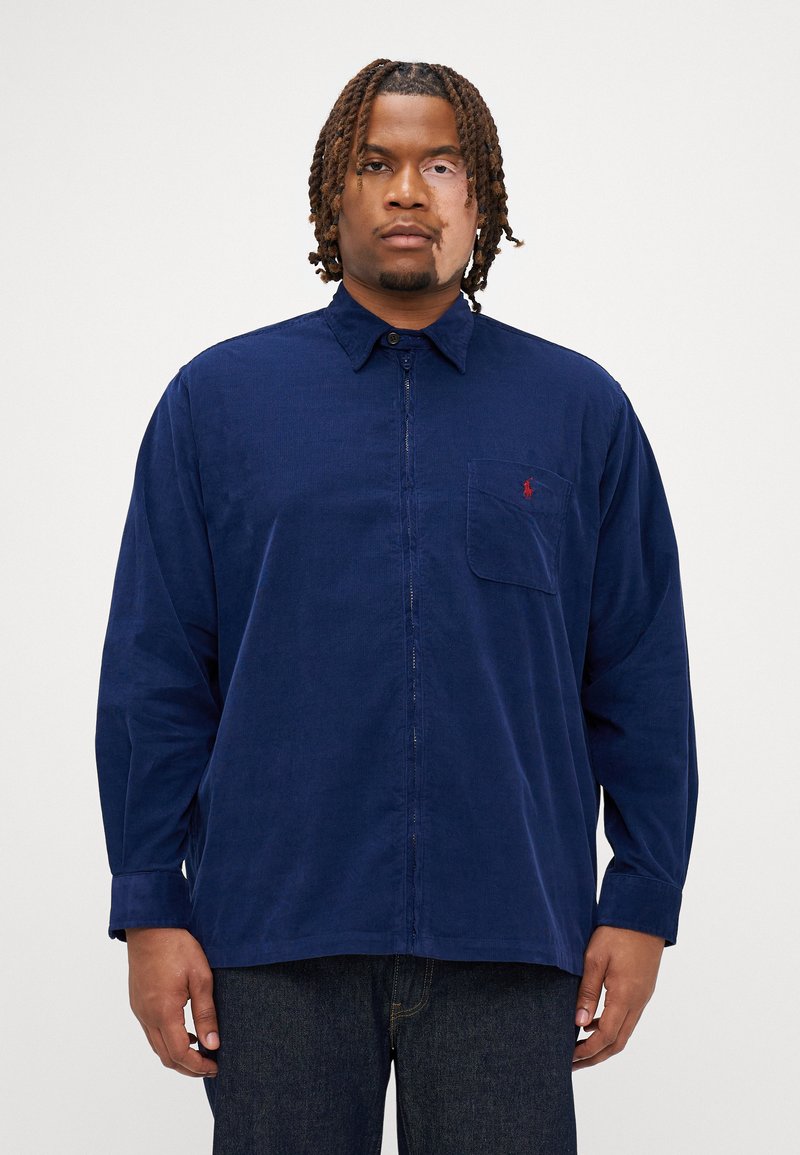 Ναυτικό καπιτονέ πουκάμισο με κουμπιά, γιακά, μονή θήκη στη θήκη, και μικρό κόκκινο λογότυπο. Μακριά μανίκια με κουμπιά στους καρπούς και ίσια γραμμή.