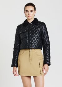 Veste en cuir matelassé noire, cropped, avec un col texturé noir et une poche frontale, associée à une mini-jupe beige avec des poches latérales.