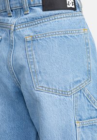 Světlemodré riflové kalhoty mají viditelné švy, zadní kapsu a černou značkovou nášivku blízko pásku. Materiál vypadá mírně drsně.