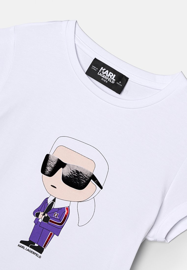 Camiseta de algodón blanca con un personaje de dibujos animados vestido con un traje púrpura, gafas de sol y detalles rojos, etiquetada "KARL LAGERFELD KIDS."