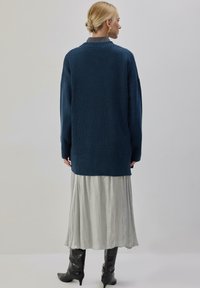 Long pull bleu tricoté à texture côtelée, associé à une jupe fluide gris clair et à des bottes noires montantes jusqu'aux genoux.