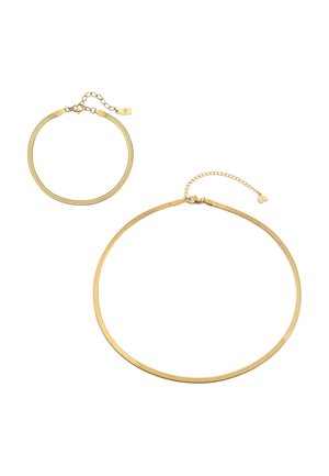 Mann im weißen Tanktop und hellgrauer Hose, der sein Kinn auf die Hände stützt und am linken Handgelenk ein dünnes goldenes Armband trägt.