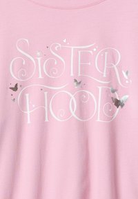 Világos rózsaszín pulóver, rajta fehér írott betűkkel a "SISTERHOOD" szó, ezüst metál pillangó díszítésekkel és dekoratív csillogó elemekkel.