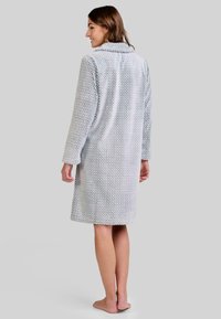 Mujer de pie descalza, vistiendo una bata de baño texturizada de color gris claro hasta las rodillas, de espaldas con la cabeza girada hacia un lado, sonriendo.