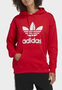 Röd huvtröja med känguruficka, som har en stor vit Adidas-logotyp och tre vita ränder på varumärkesemblemet.