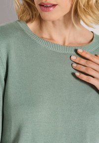 Frau in einem hellgrünen Strickpullover, die mit einer manikürten Hand den Ausschnitt berührt, während auf ihren Lippen ein natürlicher rosa Lippenstift zu sehen ist.