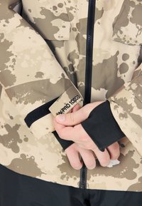 Beige takki mustilla yksityiskohdilla, jossa on vettä hylkivä kangas, abstrakti roiske-kuvio ja hihassa on etiketti, jossa lukee "WP PRO 10000".