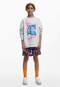 Felpa grigia con un design di un personaggio dei cartoni animati blu e testo colorato. Abbinata a una gonna nera con motivi e calzini arancioni al ginocchio.