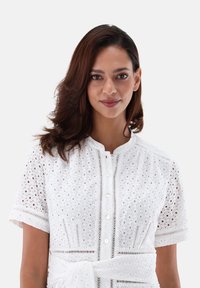 Weißes Blusenoberteil aus Baumwolle mit kurzen Ärmeln, Knopfleiste und subtiler Textur. Es hat einen gebundenen Gürtel und aufwendige Cut-out-Muster.