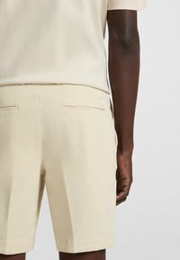 Pantaloni corti beige testurizzati con taglio sartoriale, dotati di tasche posteriori e una cintura liscia, indossati con una maglietta di un beige chiaro.