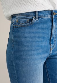 Primer plano de una persona que lleva jeans de mezclilla azules y un suéter trenzado texturizado blanco, mostrando la cintura, el bolsillo delantero y los detalles de costura.