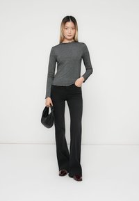 Sieviete, tērpusies pelēkā, garu piedurkņu ribveida topā, melnās izplatītās biksēs, bordo krāsas kurpēs, tur rokās mazu melnu rokas somiņu, stāvot pret baltu fonu.