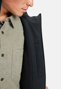 Homme portant une chemise gris clair à boutons avec une poche poitrine et une veste noire partiellement ouverte avec une fermeture éclair sur le côté droit.