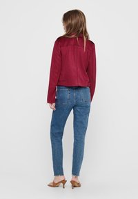 Femme debout de dos, portant une veste rouge, un jean bleu aux bords effilochés, et des sandales à talons beiges, sur un fond uni.