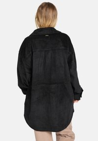 Schwarze Cordjacke mit lockerer Passform, ausgestattet mit einem Kragen und einem asymmetrischen Saum. Texturierte senkrechte Rillen sind auf dem Stoff sichtbar.