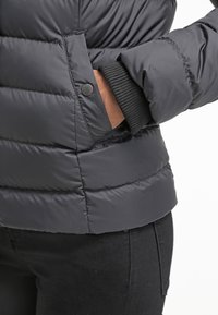 Černá péřová bunda s prošívaným vzorem, obsahující detail knoflíku a žebrované manžety, zobrazená od pasu, s viditelnou boční kapsou.