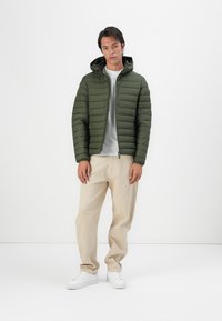 Puffer jacket in dark green with horizontal quilted pattern, a hood, and a front zipper. Paired with light beige pants and white sneakers.