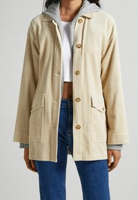 Giacca in velluto a coste con esterno beige, cappuccio grigio e bottoni in legno. Presenta due tasche frontali e una vestibilità comoda.