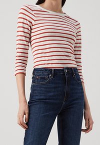 Mujer con una blusa de rayas blanca y roja de manga larga metida en unos vaqueros oscuros de cintura alta, de pie con los brazos relajados.