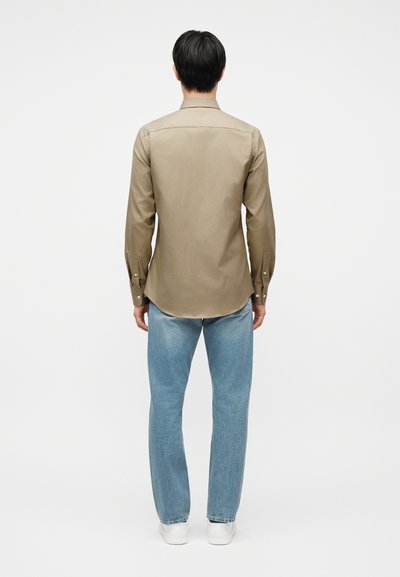 Άνδρας γυρισμένος με την πλάτη φοράει μπεζ μακρυμάνικο πουκάμισο, ανοιχτόχρωμα μπλε τζιν και λευκά αθλητικά παπούτσια μπροστά σε απλό λευκό φόντο.