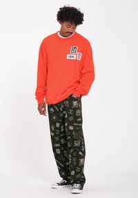 Oransje sweatshirt med frontlapper, kombinert med løstsittende grønne bukser med mønster og svarte grafiske design. Svarte sneakers fullfører antrekket.