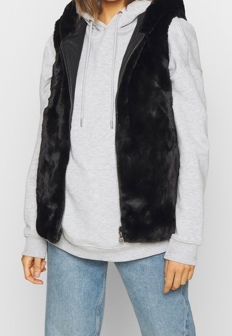 Czarna kamizelka z sztucznego futra z zapięciem na suwak i skórzanymi akcentami, noszona na szarym hoodie, uzupełniona jasnoniebieskimi dżinsami. Miękka tekstura i bezrękawnikowy krój.