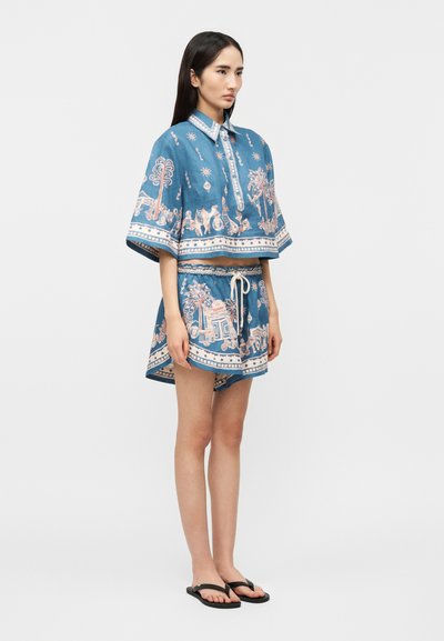 Γυναίκα με μπλε διπλό σύνολο με μοτίβα φοίνικα και ζώων, στέκεται πλαγίως με χαλαρά χέρια, μαύρα σαγιονάρες σε απλό φόντο.