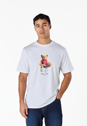 Νέος άνδρας που φοράει λευκό t-shirt με αρκουδάκι σε μπλούζα μπάσκετ, κρατώντας μια μπάλα μπάσκετ, συνδυασμένο με μπλε τζιν, στέκεται μπροστά σε λευκό φόντο.