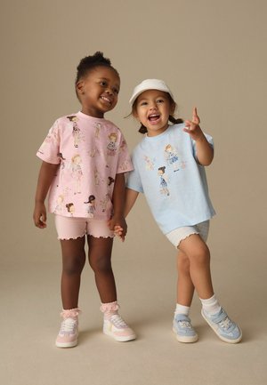 To smilende småbørn, der holder hinanden i hånden, iført trykte pastel T-shirts, shorts og sneakers, står foran en ensfarvet beige baggrund.