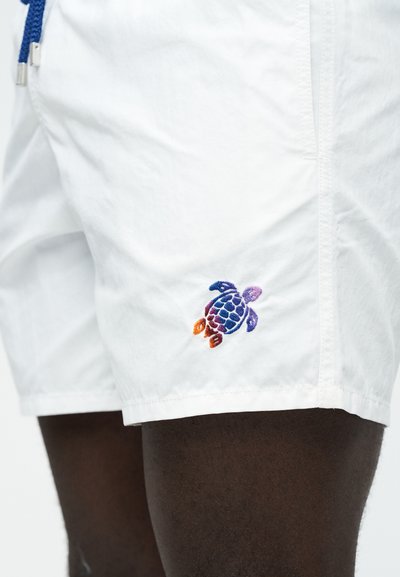 Λευκά σορτς με μπλε κορδόνι που φοριούνται σε σκουρόχρωμο πόδι, με μικρή κεντητή πολύχρωμη θαλάσσια χελώνα κοντά στο τελείωμα.