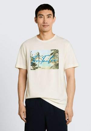 Mężczyzna w kremowym T-shircie z tropikalnym nadrukiem plaży i niebieskim napisem, w połączeniu z czarnymi spodniami, stoi na jasnym, jednolitym tle.