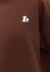 Tissu marron avec une petite lettre blanche stylisée "L" imprimée sur la partie supérieure de la poitrine.
