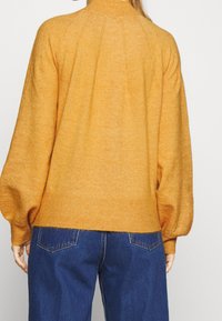 Person wearing a mustard yellow knit sweater with puffed long sleeves and blue denim jeans, shown from the back against a white background.