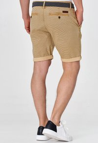 Shorts rayés couleur cannelle avec revers retroussés, chaussures contrastantes noires et blanches, et ceinture à motifs. Deux poches arrière et détails cousus visibles.