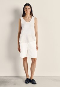 Kobieta o długich ciemnych włosach, ubrana w bezrękawnik w kolorze białym, białe szorty do kolan oraz czarne mokasyny, stoi na jednolitym tle.