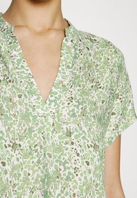 Blusa verde a fantasia con scollatura a V, maniche corte e vestibilità ampia. Il tessuto è leggero, morbido al tatto e con una leggera lucentezza.